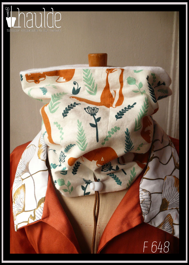 Tour de cou en tissu blanc imprimé renard roux et végétaux clairsemés dans les tons verts, le dessin est de style naïf, les renards sont dans plusieurs postures, devant il apparait couché, mais il y en a aussi des assis Cordon de serrage marron avec un stop blanc Vu relevé, sur un mannequin de couture