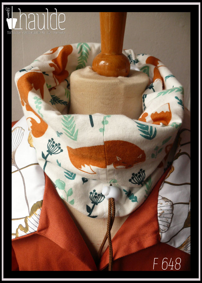 Tour de cou en tissu blanc imprimé renard roux et végétaux clairsemés dans les tons verts, le dessin est de style naïf, les renards sont dans plusieurs postures, devant il apparait couché, mais il y en a aussi des assis Cordon de serrage marron avec un stop blanc Vu sur un mannequin de couture