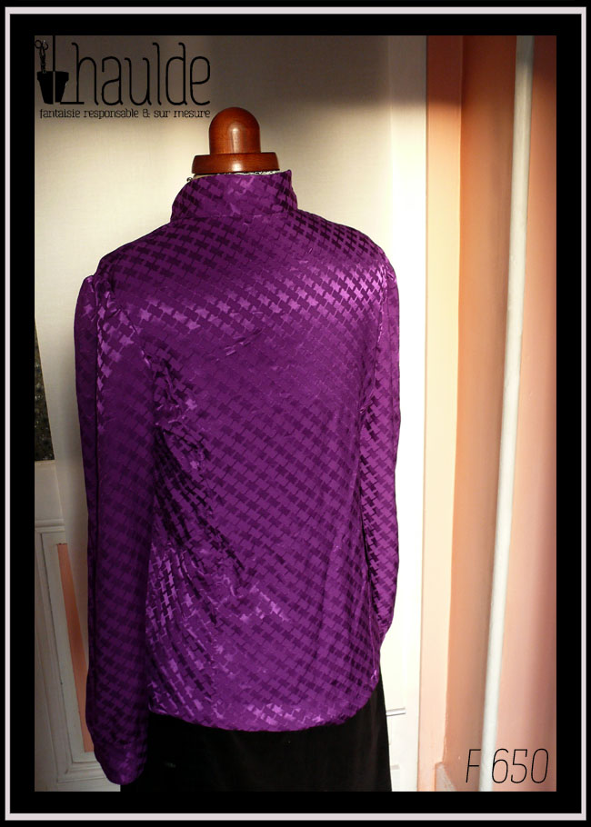 Chemisier à manches longues, col officier incliné vers le centre (peut se porter cassé) en jacquard de viscose, motif rectangles ondulés reliés par des lignes, comme le motif traditionnel dit "pied de poule" Légères fronces horizontales au niveau de la poitrine, vu de dos sur un mannequin de couture