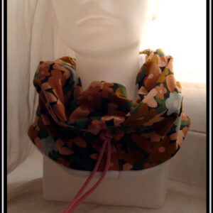 Tour de cou en jersey gratté imprimé, fleurs ocre, corail, écru et bleu pâle sur fond noir avec feuilles vertes stylisées. Cordon de serrage rose bonbon, stop de la même couleur, clochette couleur acier pour recouvrir les noeuds Vu de face sur une tête de mannequin