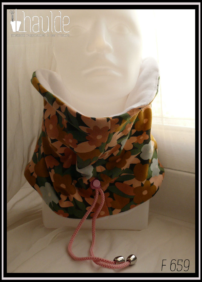 Tour de cou en jersey gratté imprimé, fleurs ocre, corail, écru et bleu pâle sur fond noir avec feuilles vertes stylisées. Cordon de serrage rose bonbon, stop de la même couleur, clochette couleur acier pour recouvrir les noeuds Vu relevé sur une tête de mannequin