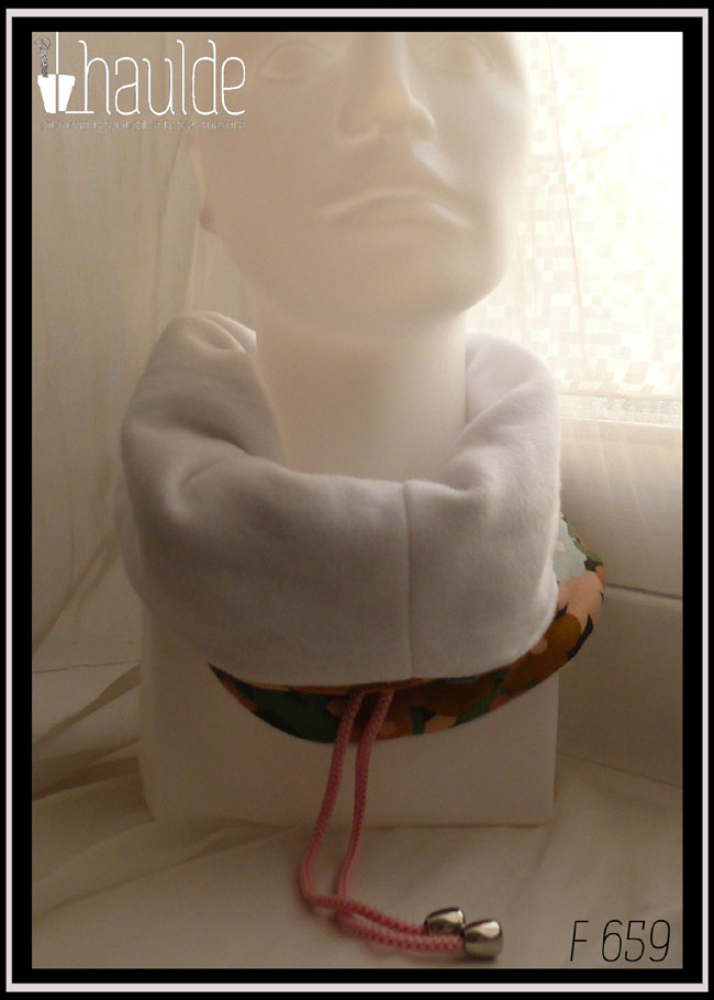 Tour de cou en jersey gratté imprimé, fleurs ocre, corail, écru et bleu pâle sur fond noir avec feuilles vertes stylisées. Cordon de serrage rose bonbon, stop de la même couleur, clochette couleur acier pour recouvrir les noeuds Vu replié côté doublure en polaire blanche sur une tête de mannequin