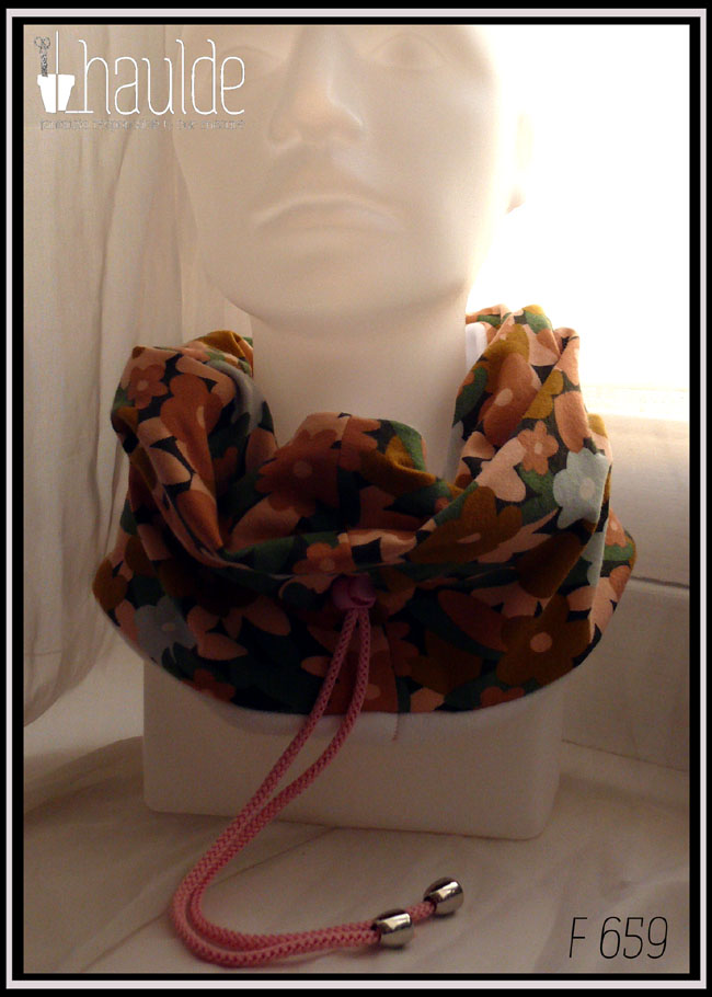 Tour de cou en jersey gratté imprimé, fleurs ocre, corail, écru et bleu pâle sur fond noir avec feuilles vertes stylisées. Cordon de serrage rose bonbon, stop de la même couleur, clochette couleur acier pour recouvrir les noeuds Vu de face sur une tête de mannequin