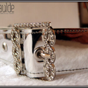 Une ceinture en simili cuir argenté rutilant avec une boucle rectangulaire décorée de strass Vue en gros plan de la boucle, les strass forment un genre de tresse ouverte de chaque côté dans le sens de la hauteur