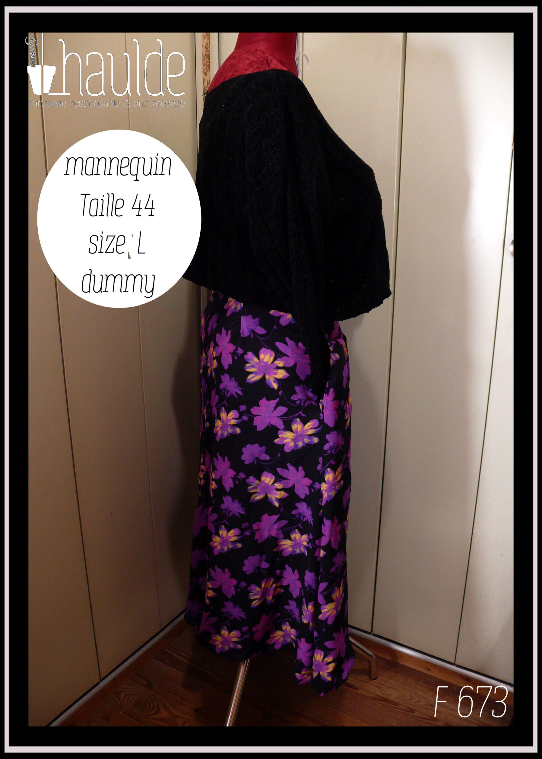 Jupe asymétrique en crêpe noir imprimé fleurs stylisées violet électrique, certaines ont des touches de jaune pâle ou de fuchsia fluo La jupe est très fendue et plus courte côté gauche, longue et très plissée dans le dos et côté droit. Vue de profil côté droit (long) sur un mannequin de couture taille 44 avec un pull noir