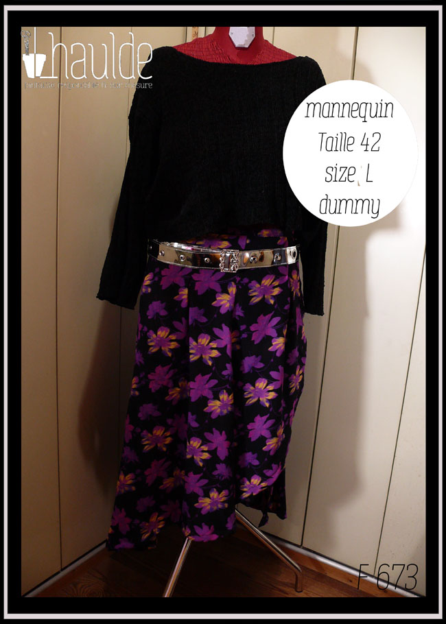 Jupe asymétrique en crêpe noir imprimé fleurs stylisées violet électrique, certaines ont des touches de jaune pâle ou de fuchsia fluo La jupe est très fendue et plus courte côté gauche, longue et très plissée dans le dos et côté droit. Vue de face sur un mannequin de couture taille 42 avec une ceinture argentée brillante et un pull noir