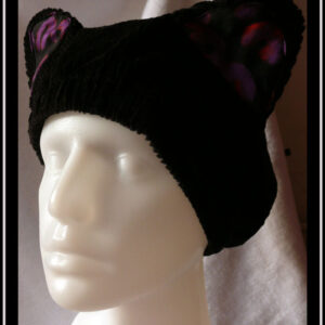 Bonnet en velours côtelé noir avec deux oreilles de chats dont l'intérieur est garni de tissu noir imprimé motifs panthère stylisé violet légèrement soulignés de orange Vu sur une tête de mannequin de trois quarts face