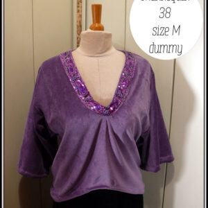 Haut col en V plongeant, manches kimono trois quarts en velours mauve. L'encolure est garnie d'un galon brodé de sequins violets et fuchsia qui forment des motifs paisley Vu sur un mannequin de couture taille 38