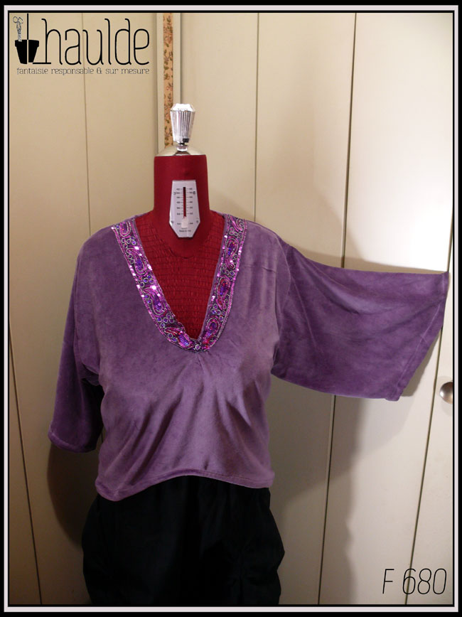 Haut col en V plongeant, manches kimono trois quarts en velours mauve. L'encolure est garnie d'un galon brodé de sequins violets et fuchsia qui forment des motifs paisley La manche gauche est suspendu en l'air pour qu'on voit son amplitude