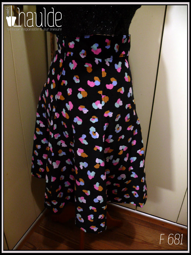 Jupe à ceinture large, pans évasés en velours milleraies noir imprimé de taches regroupées par trois ou quatre, mauve, vert pâle, ocre, rose et bleu, qui pourraient faire penser à un pelage animal (le motif s'appelle animal skin) Vue de profil sur un mannequin de couture