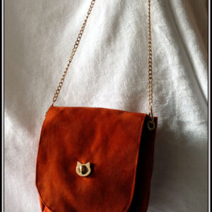 Sac à bandoulière à rabat en suédine orange foncé, bandoulière chaine et fermoir tourniquet en forme de tête de chat en acier doré. Vu suspendu. Le rabat a les coins arrondis