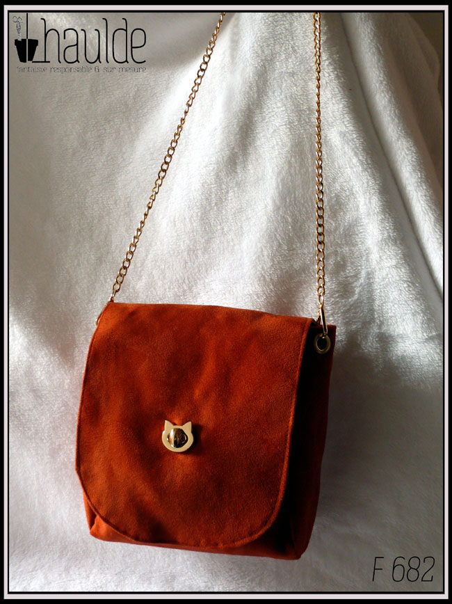 Sac à bandoulière à rabat en suédine orange foncé, bandoulière chaine et fermoir tourniquet en forme de tête de chat en acier doré. Vu suspendu. Le rabat a les coins arrondis