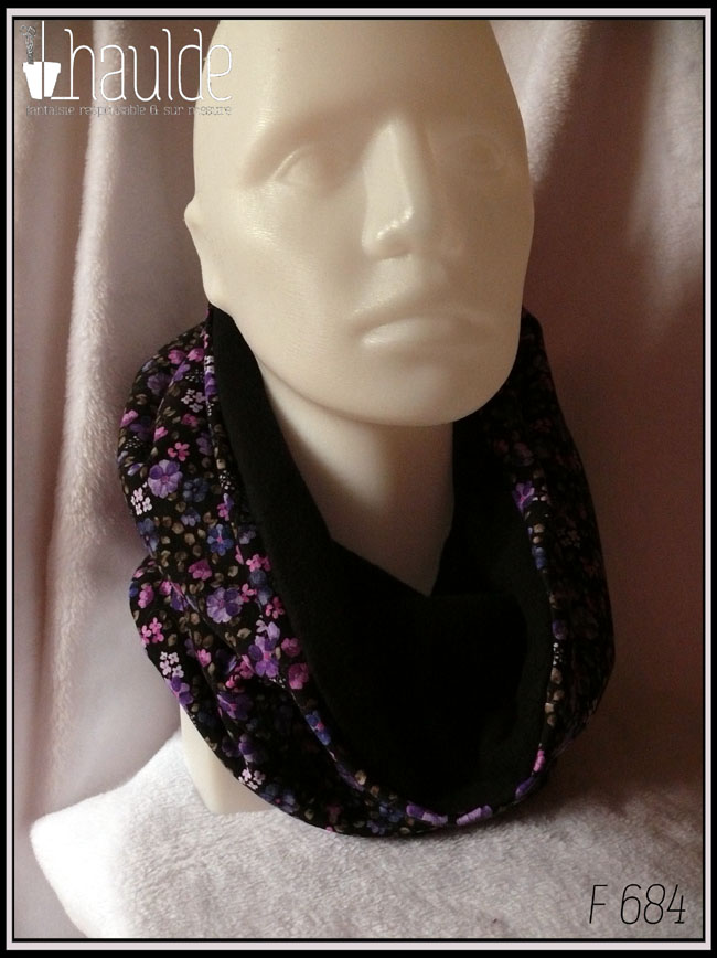 Tour de cou ample qui peut se porter en capuche, en jersey noir imprimé petites fleurs réalistes dans les tons violets doublé de polaire de coton noire Vu porté comme un tour de cou classique sur une tête de mannequin de trois quarts face