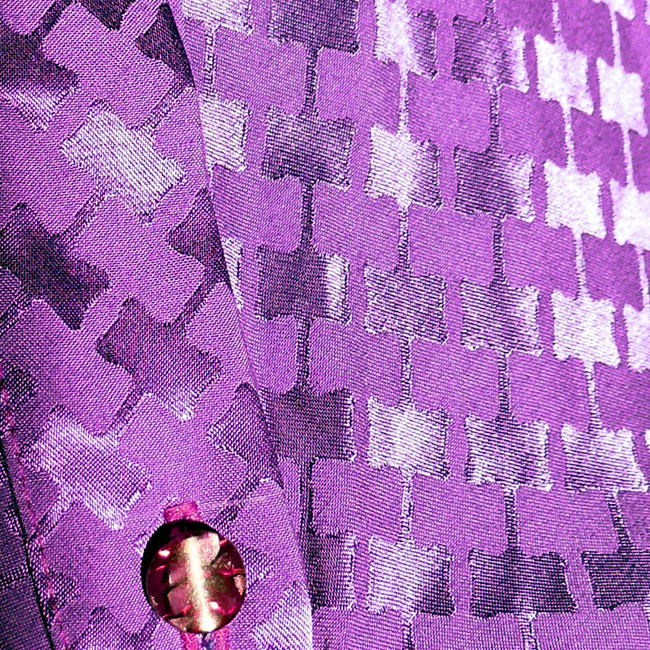 gros plan du jacquard de viscose violet, motif rectangles ondulés reliés par des lignes, presque comme le motif traditionnel dit "pied de poule" On aperçoit également un des boutons du chemisier, pourpre avec des paillettes incrustées