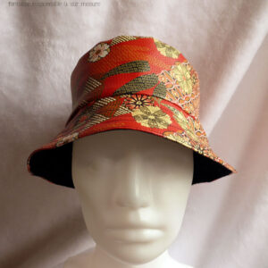 Bob en simili cuir rouge imprimé motifs floraux d'esthétique japonaise dans les tons kakis, jaune pâle et orange, cernés de noir. L'autre face du bob est en velours milleraies noir Vu sur une tête de mannequin