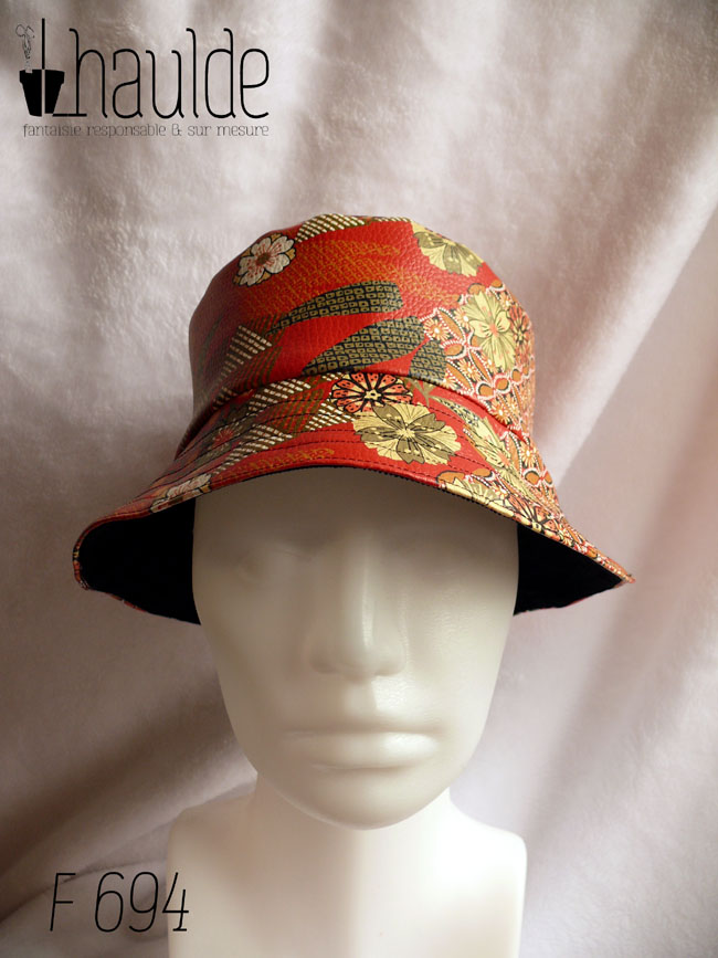 Bob en simili cuir rouge imprimé motifs floraux d'esthétique japonaise dans les tons kakis, jaune pâle et orange, cernés de noir. L'autre face du bob est en velours milleraies noir Vu sur une tête de mannequin