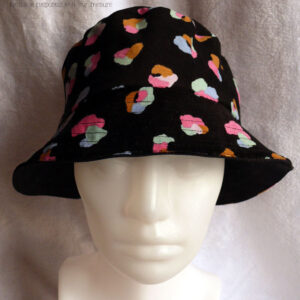 Bob en milleraies noir imprimé de taches regroupées par trois ou quatre, mauve, vert pâle, ocre, rose et bleu, qui pourraient faire penser à un pelage animal (le motif s'appelle animal skin) Réversible sur du sergé noir uni Vu de dessus côté milleraies, porté par une tête de mannequin