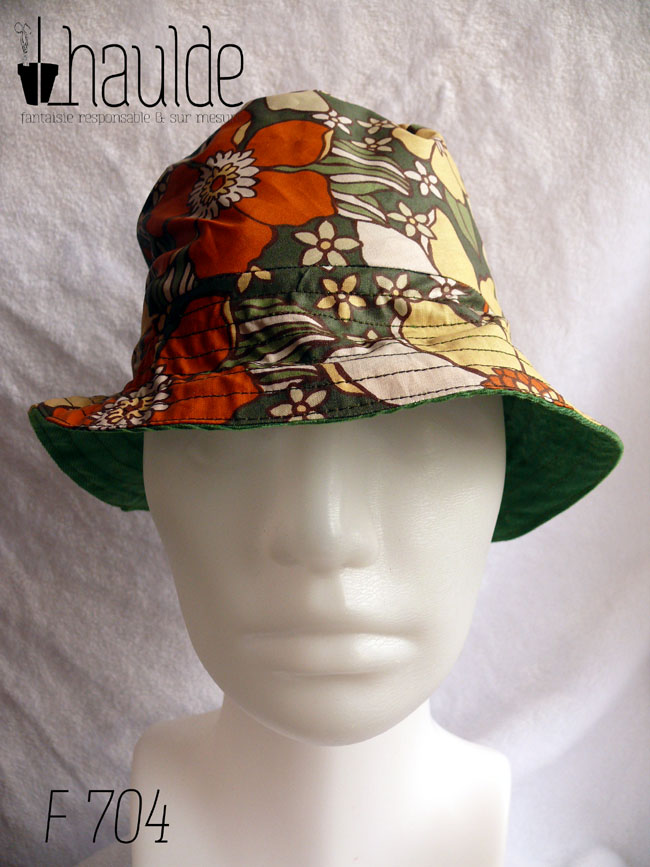 Bob réversible, vu du côté en toile enduite, fond kaki imprimé de grandes fleurs oranges et jaune pâle dans le style des années 70. La tête de mannequin est un peu trop grande pour ce chapeau pour enfant
