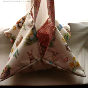 Sac à tarte, le dessus est en toile blanche imprimée de petits lapins, poules couveuses, poussins, petit agneau endormis, fleurs variées, et nids d'oeufs de Pâques dans un style illustration jeunesse à l'aquarelle. L'intérieur est en tissus spécial contact alimentaire blanc imprimé de carrés oranges à intérieur blanc alignés avec de temps en temps un carré plein mauve, rose, jaune ou orange Le sac est vu de face, les anses tenues en l'air.