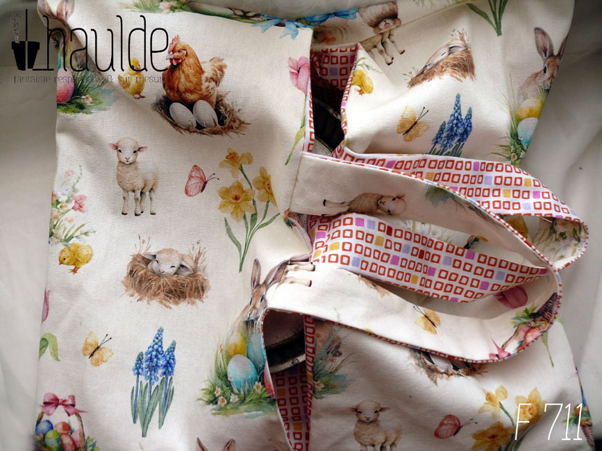 Sac à tarte, le dessus est en toile blanche imprimée de petits lapins, poules couveuses, poussins, petit agneau endormis, fleurs variées, et nids d'oeufs de Pâques dans un style illustration jeunesse à l'aquarelle. L'intérieur est en tissus spécial contact alimentaire blanc imprimé de carrés oranges à intérieur blanc alignés avec de temps en temps un carré plein mauve, rose, jaune ou orange Vu de dessus