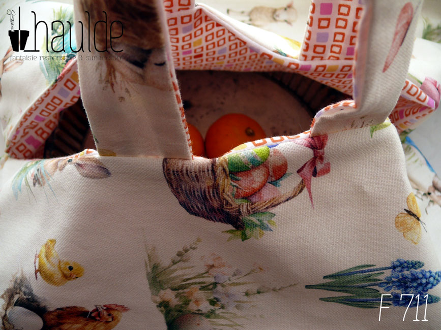 Vue en gros plan du sac, anses levée, à l'intérieur on devine un plat à tarte en inox avec des citrons bergamotes posés dessus Sac à tarte, le dessus est en toile blanche imprimée de petits lapins, poules couveuses, poussins, petit agneau endormis, fleurs variées, et nids d'oeufs de Pâques dans un style illustration jeunesse à l'aquarelle. L'intérieur est en tissus spécial contact alimentaire blanc imprimé de carrés oranges à intérieur blanc alignés avec de temps en temps un carré plein mauve, rose, jaune ou orange