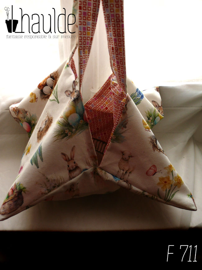 Sac à tarte, le dessus est en toile blanche imprimée de petits lapins, poules couveuses, poussins, petit agneau endormis, fleurs variées, et nids d'oeufs de Pâques dans un style illustration jeunesse à l'aquarelle. L'intérieur est en tissus spécial contact alimentaire blanc imprimé de carrés oranges à intérieur blanc alignés avec de temps en temps un carré plein mauve, rose, jaune ou orange Le sac est vu de face, les anses tenues en l'air.