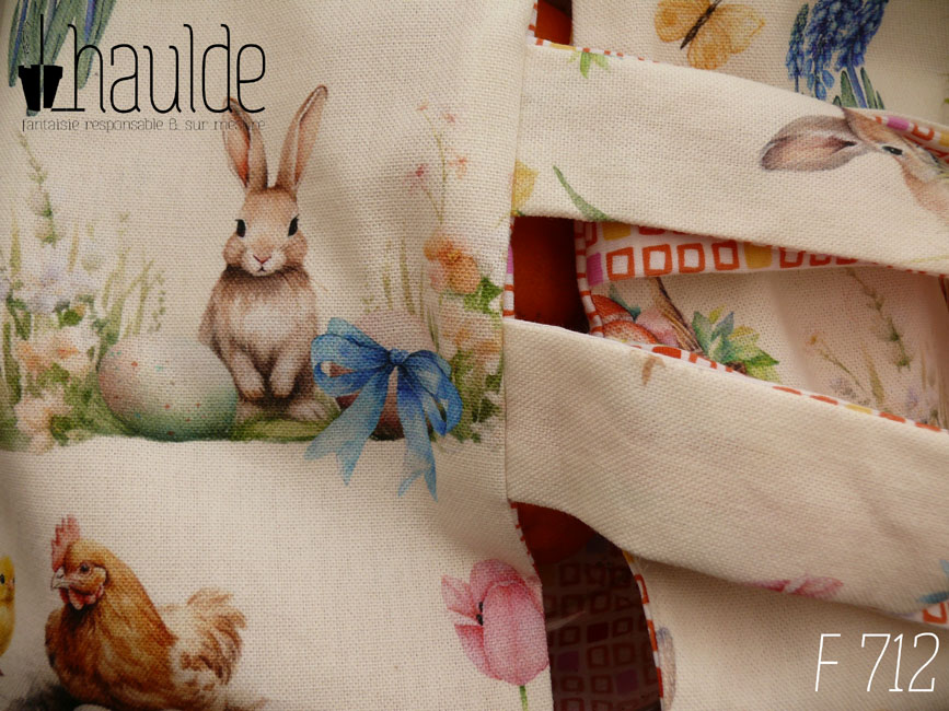 Vue en gros plan du sac, de dessus. Sac à tarte, le dessus est en toile blanche imprimée de petits lapins, poules couveuses, poussins, petit agneau endormis, fleurs variées, et nids d'oeufs de Pâques dans un style illustration jeunesse à l'aquarelle. L'intérieur est en tissus spécial contact alimentaire blanc imprimé de carrés oranges à intérieur blanc alignés avec de temps en temps un carré plein mauve, rose, jaune ou orange