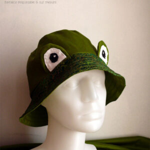 Bob réversible en sergé vert moyen assez lumineux, et des yeux de grenouille (tour vert, centre blanc avec la pupille noire et un petit point blanc) dressés au-dessus du rebord en tissu imprimé, fond vert foncé, feuillage vert vif souligné de doré. Vu de trois quarts face sur un mannequin de couture