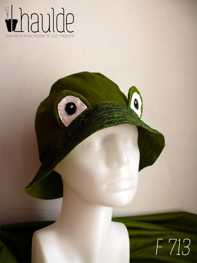 Bob réversible en sergé vert moyen assez lumineux, et des yeux de grenouille (tour vert, centre blanc avec la pupille noire et un petit point blanc) dressés au-dessus du rebord en tissu imprimé, fond vert foncé, feuillage vert vif souligné de doré. Vu de trois quarts face sur un mannequin de couture