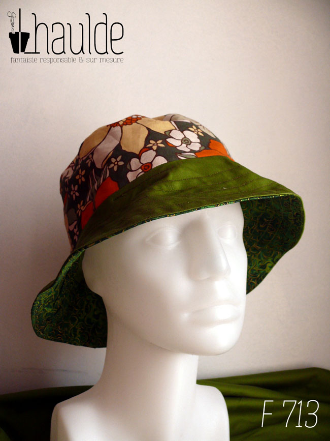 Bob réversible en sergé vert moyen assez lumineux, avec de la toile enduite imprimée fleurs façon années 70 dans les tons blancs, oranges et jaune pâle sur donc kaki foncé et tissu imprimé, fond vert foncé, feuillage vert vif souligné de doré. Ici la bande centrale est en toile enduite, le dessus du rebord en sergé, le dessous en tissu vert imprimé Vu de trois quarts face sur une tête de mannequin