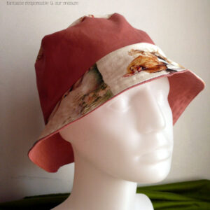 Bob réversible en toile blanche imprimée de petits lapins, poules couveuses, poussins, petits agneaux endormi dans un nid ou debout, fleurs variées, et nids d'oeufs de Pâques dans un style illustration jeunesse à l'aquarelle et sergé rose tendre Ici la bande centrale est rose, le dessus du bord en imprimé, le dessous du bord est rose. Vu sur une tête de mannequin, de trois quarts face