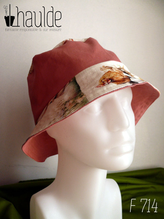 Bob réversible en toile blanche imprimée de petits lapins, poules couveuses, poussins, petits agneaux endormi dans un nid ou debout, fleurs variées, et nids d'oeufs de Pâques dans un style illustration jeunesse à l'aquarelle et sergé rose tendre Ici la bande centrale est rose, le dessus du bord en imprimé, le dessous du bord est rose. Vu sur une tête de mannequin, de trois quarts face