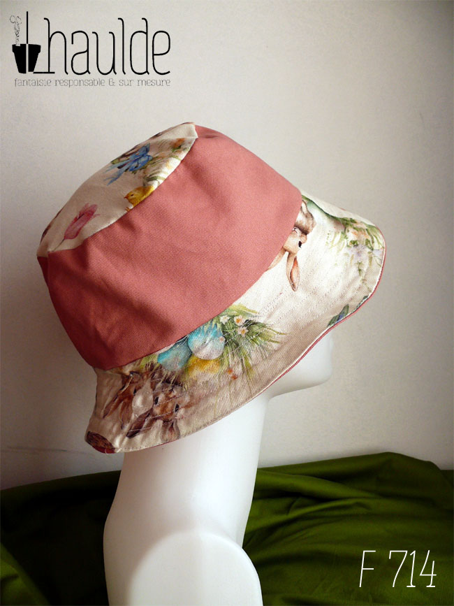 Bob réversible en toile blanche imprimée de petits lapins, poules couveuses, poussins, petits agneaux endormi dans un nid ou debout, fleurs variées, et nids d'oeufs de Pâques dans un style illustration jeunesse à l'aquarelle et sergé rose tendre Ici la bande centrale est rose, le dessus du bord en imprimé, le dessus du bob est également en imprimé Vu sur une tête de mannequin, de profil