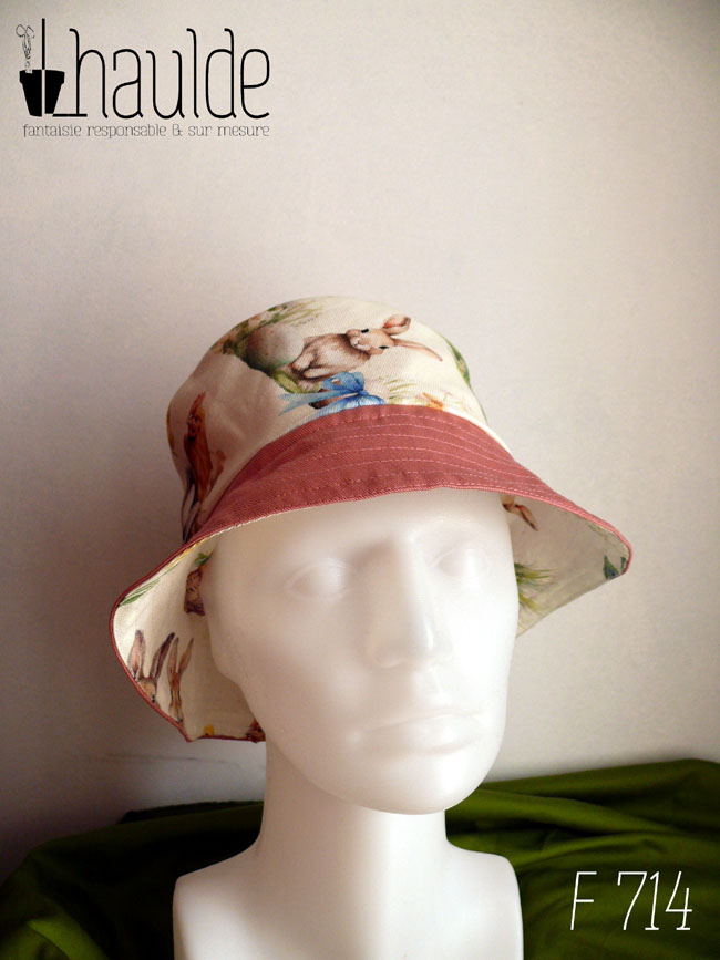 Bob réversible en toile blanche imprimée de petits lapins, poules couveuses, poussins, petits agneaux endormi dans un nid ou debout, fleurs variées, et nids d'oeufs de Pâques dans un style illustration jeunesse à l'aquarelle et sergé rose tendre Ici la bande centrale est en imprimé, le dessus du bord rose, le dessous du bord en imprimé Vu sur une tête de mannequin, de face