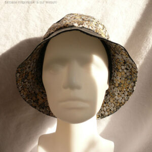 Bob en simili cuir noir et tissu imprimé. Les motifs sont des ovales concentriques de petite taille inspirés par Klimt dans les tons gris rehaussés de doré. Ici le dessus du bob est en uni, la bande centrale en imprimé, le bord en uni Vu de face sur une tête de mannequin, le dessous du bord en imprimé est bien visible