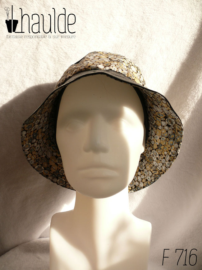 Bob en simili cuir noir et tissu imprimé. Les motifs sont des ovales concentriques de petite taille inspirés par Klimt dans les tons gris rehaussés de doré. Ici le dessus du bob est en uni, la bande centrale en imprimé, le bord en uni Vu de face sur une tête de mannequin, le dessous du bord en imprimé est bien visible