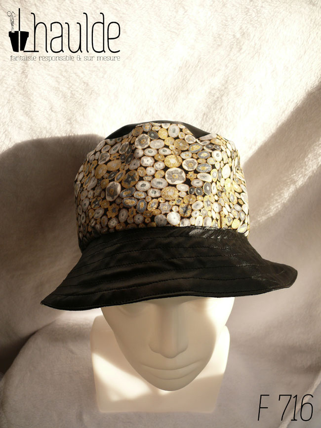 Bob en simili cuir noir et tissu imprimé. Les motifs sont des ovales concentriques de petite taille inspirés par Klimt dans les tons gris rehaussés de doré. Ici le dessus du bob est en uni, la bande centrale en imprimé, le bord en uni Vu de dessus sur une tête de mannequin