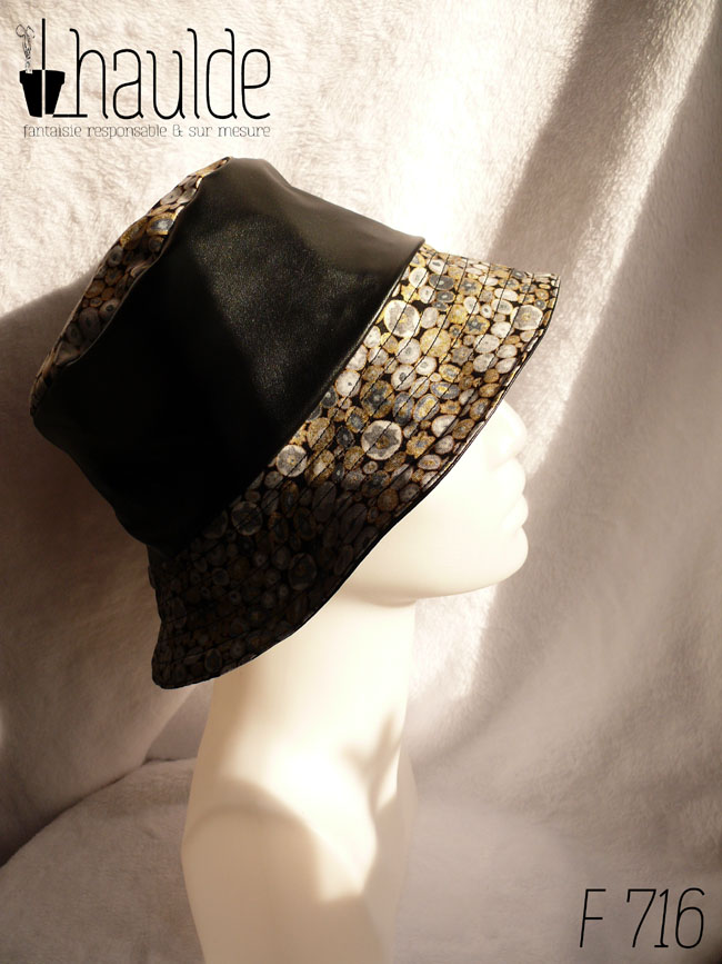 Bob en simili cuir noir et tissu imprimé. Les motifs sont des ovales concentriques de petite taille inspirés par Klimt dans les tons gris rehaussés de doré. Ici le dessus du bob est en imprimé, la bande centrale en uni, le bord en imprimé Vu de profil sur une tête de mannequin