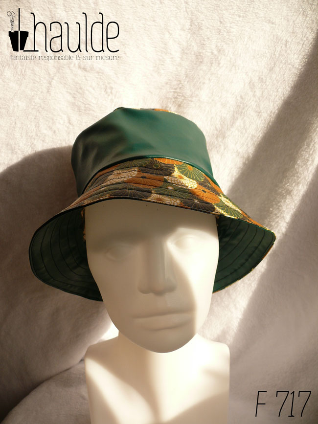 Bob en simili cuir vert et toile enduite imprimée de fleurs stylisées de type chrysanthèmes alignées les unes sur les autres comme un motif queue de paon. Elles sont blanches ou marron roux, ou noires ou vertes. Ici le dessus du bob est en imprimé, la bande centrale en uni, le bord en imprimé Vu de face sur une tête de mannequin