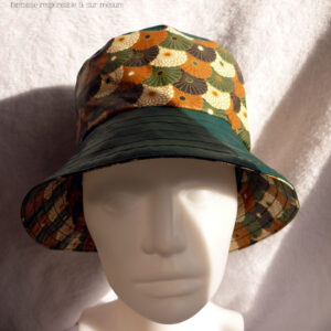 Bob en simili cuir vert et toile enduite imprimée de fleurs stylisées de type chrysanthèmes alignées les unes sur les autres comme un motif queue de paon. Elles sont blanches ou marron roux, ou noires ou vertes. Ici le dessus du bob est en simili uni, la bande centrale en imprimé, le bord en uni Vu de face sur une tête de mannequin