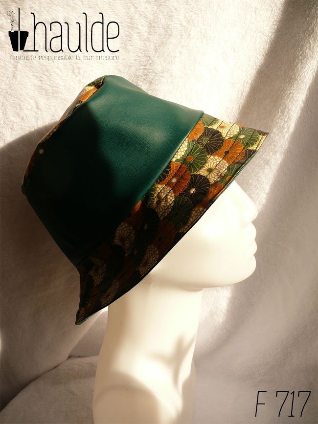 Bob en simili cuir vert et toile enduite imprimée de fleurs stylisées de type chrysanthèmes alignées les unes sur les autres comme un motif queue de paon. Elles sont blanches ou marron roux, ou noires ou vertes. Ici le dessus du bob est en imprimé, la bande centrale en uni, le bord en imprimé Vu de profil sur une tête de mannequin