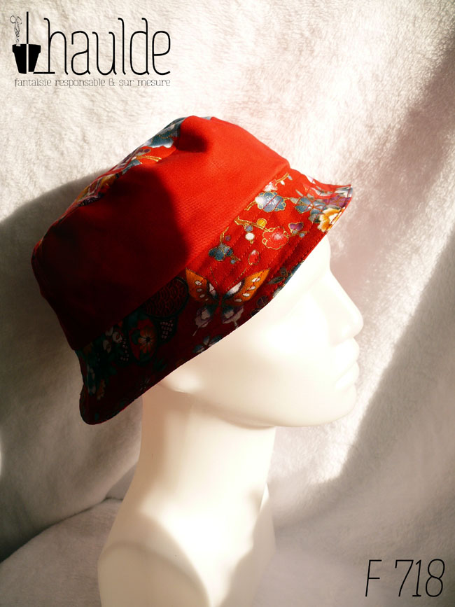 Bob court en tissu rouge imprimé pivoines, papillons et autres fleurs multicolores soulignées de doré, d'inspiration Est asiatique (Chine ou Japon) et sergé rouge, réversible. Ici le dessus est en tissu imprimé, la bande centrale en rouge uni, le bord en imprimé vu de profil sur une tête de mannequin