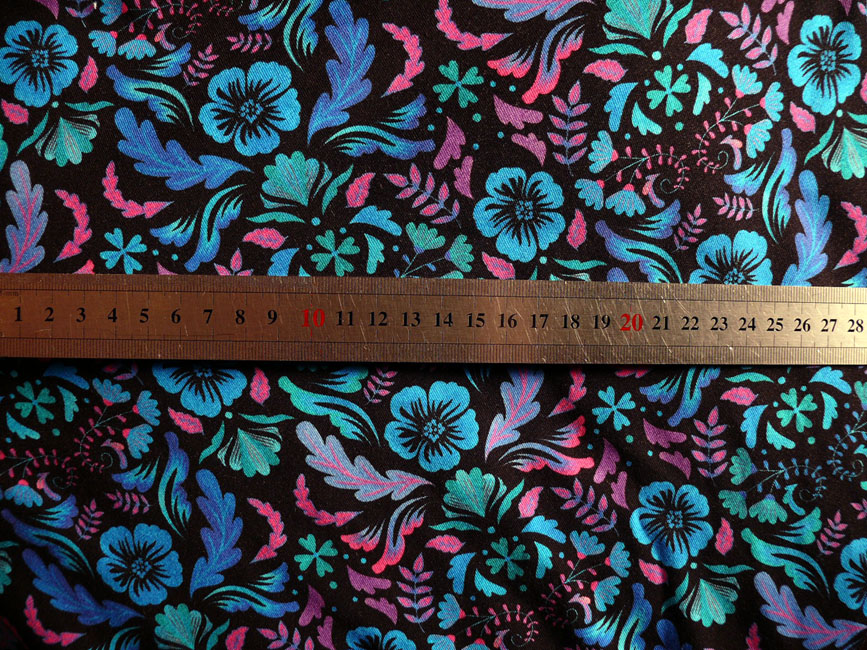 jersey noir imprimé fleurs et végétaux stylisés répartis de manière symétriques, dans les tons bleus fluo, vert émeraude fluo, rose et mauve fluo Le mètre posé dessus indique que les fleurs font 2.5 cm de large max, les feuilles sont longues de 4 cm max