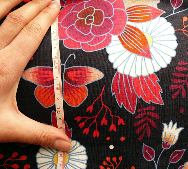 jersey noir imprimé fleurs et papillons stylisés dans les tons rouges avec quelques fleurs blanches à coeur jaunes de types marguerites, tous cernés de bleu pâle Le mètre et ma main posés dessus indiquent que les fleurs font 6 cm de large max