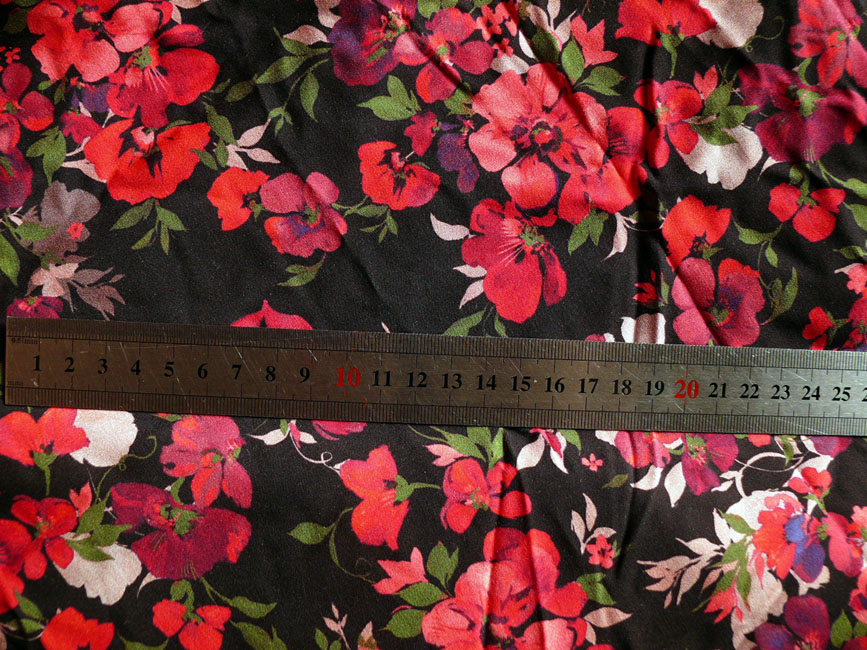 tissu souple et léger, fond noir, motifs petites fleurs rouges-roses vifs de type aubépine, avec de petites feuilles vertes et quelques "ombres" blanches pour rehausser Le mètre posé dessus indique que les motifs font 4 cm max de large