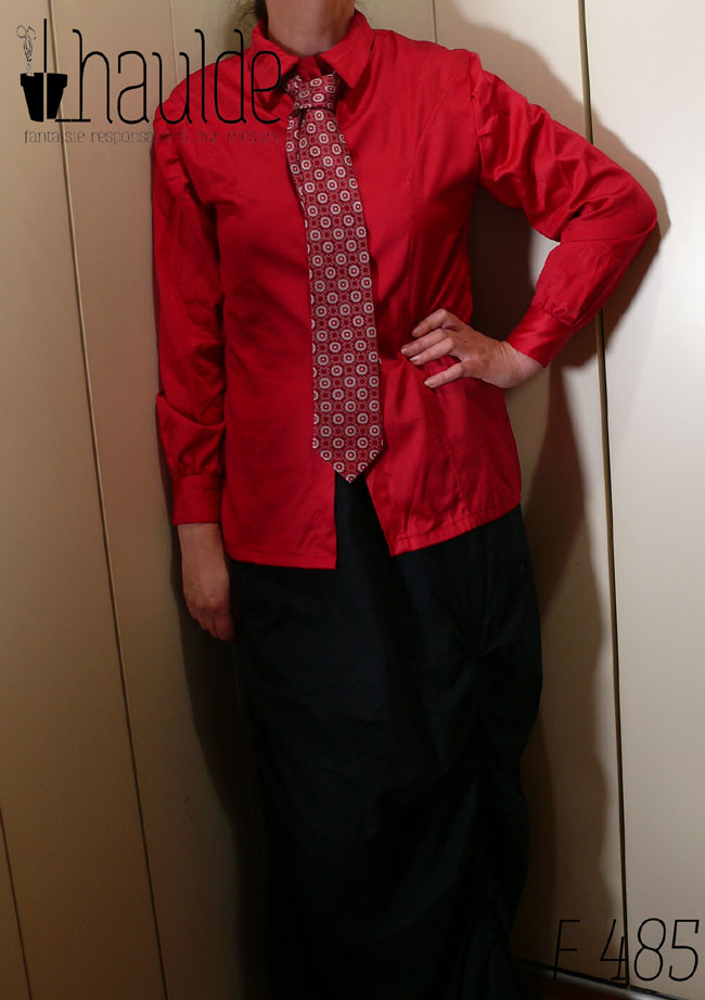 Femme debout, tête et pieds hors champ vêtue d'un chemisier manches longues en satin de coton rouge (assez mat, peu soyeux) d'une cravate rouge foncé imprimée de motifs géométriques gris perle et rouge très foncé et d'une jupe longue noire La main gauche est campée sur la taille.