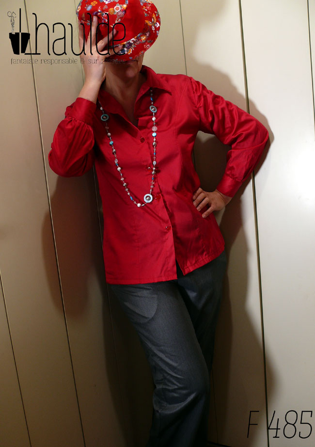 Femme debout, pieds hors champ, vêtue d'un chemisier manches longues en satin de coton rouge (assez mat, peu soyeux) Elle porte également un long collier en verre bleu et nacre blanc, un pantalon gris en laine satinée et cache sa tête avec un bob court en tissu rouge uni et tissu rouge imprimé de motifs floraux multicolore d'inspiration chinoise. La main droite tient le chapeau, la main gauche est campée sur la taille, la jambe droite se replie légèrement devant la gauche