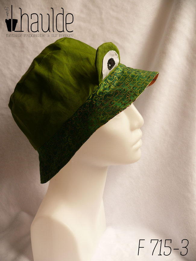 Bob réversible en sergé vert moyen assez lumineux, et des yeux de grenouille (tour vert, centre blanc avec la pupille noire et un petit point blanc) dressés au-dessus du rebord en tissu imprimé, fond vert foncé, feuillage vert vif souligné de doré. Vu de profil sur une tête de mannequin