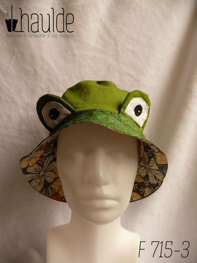 Bob réversible en sergé vert moyen assez lumineux, et des yeux de grenouille (tour vert, centre blanc avec la pupille noire et un petit point blanc) dressés au-dessus du rebord en tissu imprimé, fond vert foncé, feuillage vert vif souligné de doré. Vu de face sur une tête de mannequin