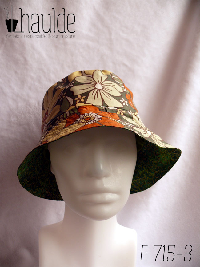 Bob réversible, vu du côté en toile enduite, fond kaki imprimé de grandes fleurs oranges et jaune pâle dans le style des années 70. Vu de face sur une tête de mannequin